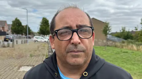 BBC A man in a black hooded top, with a blue t-shirt underneath with black glasses looks straight at the camera. He is stood on a street with a house and cars in the background.