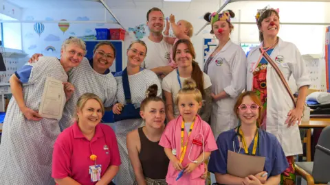 West Suffolk Hospital A group photo of men and women - some wearing patient hospital scrubs - smiling and looking into the camera while standing in a hospital ward. In the middle at the bottom is a young girl wearing pink doctors scrubs. 