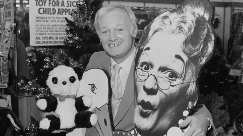 Getty Images English actor John Inman takes part in the Woolworths Toy for a Sick Child Appeal at Christmas, London, UK, 3rd December 1981. He is holding a picture of himself as a pantomime dame in the production of 'Mother Goose', in which he is appearing at the Victoria Palace Theatre. The toys will be given to children at the Great Ormond Street Hospital.