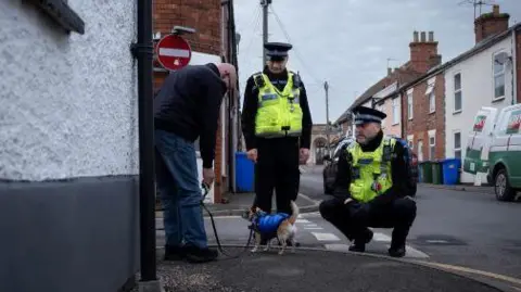 A man with a blue jacket and blue jeans talking to two police officers. The man has a small dog, which is wearing a blue coat. The officers are wearing hi-vis jackets. One officer is crouched down and looking at the dog. They are on a residential street. On the right is a row of terrace houses, with green and blue bins on the pavement. A green and white van is parked with its back door open.