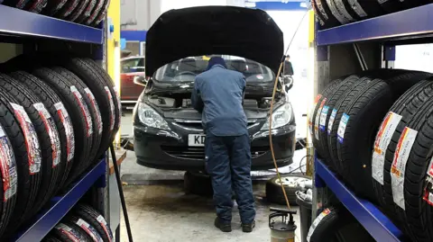 Getty Images Generic image of a mechanic checking a car's engine under the bonnet inside a garage. Shelves of tyres can be seeen on both sides.