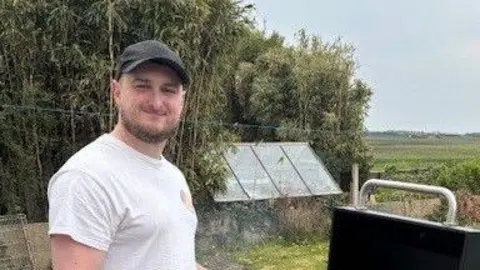 Jake Carroll smiling for a photo while making an outdoor barbecue in the countryside. He is wearing a dark baseball cap and white vest top. A garden shed and trees can be seen in the background.