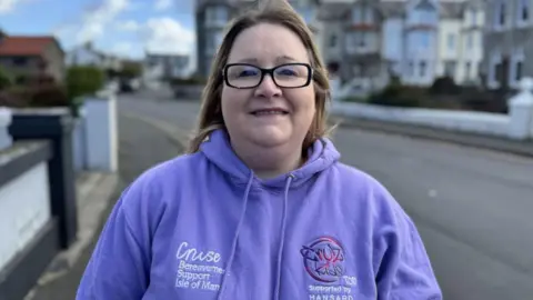 Claire is wearing a purple hoodie with Cruse Bereavement Support Isle of Man branding on it. She has shoulder length dark blonde hair blown back by the wind, and is wearing black-rimmed eye glasses.