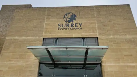 Front entrance of Surrey County Council headquarters at Woodhatch Place in Reigate. A sandy brick building with the words 'Surrey County Council' on it and the council's logo. There is also a glass door.