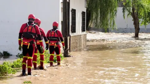 EPA Three rescue workers wearing red and yellow wetsuits with red hard hats walk through ankle-deep brown water on a Spanish street.