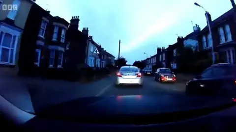 A still photo from a police pursuit video taken from the front window of the police car. A silver car can be seen driving through a housing estate at nighttime.