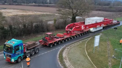 An abnormal load being escorted on a red trailer. It is travelling along a single carriageway road.