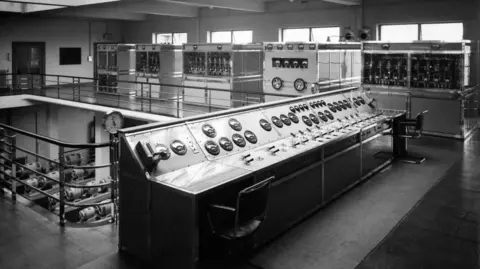 A black and white photo of control desks with dials and levers, as well as electrical boxes.