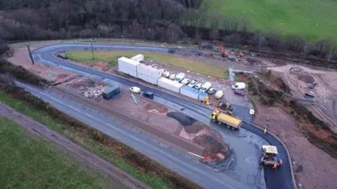 An aerial view of the construction site - a road winds around an area filled with piles of earth and grit, a series of large, white containers and lots of vehicles.