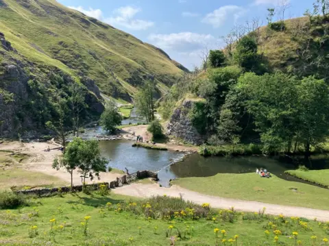 A river with green hills in the sunshine. People can be seen crossing the river over stones in the water.