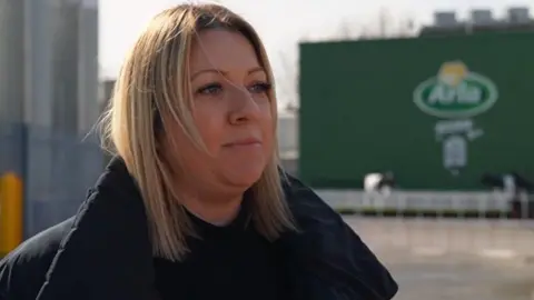 A woman with blonde hair and a dark coat stands outside the green Arla factory building.