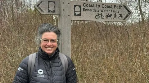 Jo Willmott is a woman in her fifties with greying hair swept back and glasses. She is wearing a black coat and is standing in front of a sign which says "Coast to Coast Ennerdale Water 1 mile".