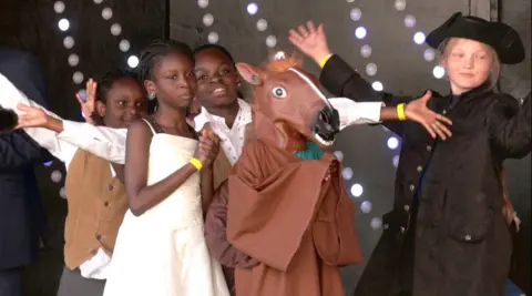 Four children on stage performing. Two boys and two girls. One girl is at the front in a white dress, her hands are clasped together. The three behind her have their arms wide open and another person is dressed in a brown horse costume.