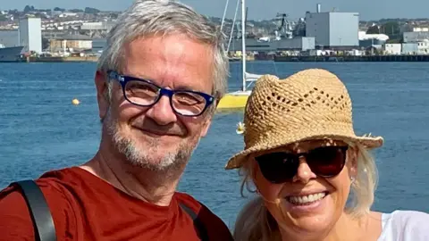 A man in a brown top with grey beard and hair with glasses and a woman in a straw hat wearing sunglasses in front of the sea.