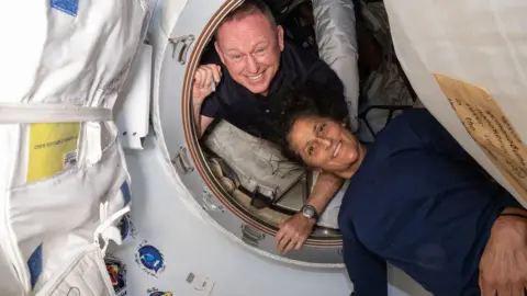 Nasa Butch Wilmore olhando através de uma escotilha na estação espacial internacional e Suni Williams deitado do lado de fora. Ambos estão sorrindo e o cabelo de Suni está acenando no ambiente de gravidade zero