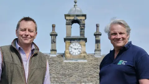 Howardian Hills/Tony Bartholomew Two men standing in front of an ornate clock tower. The man on the left is smiling and has short hair and is wearing a brown gilet and purple checked shirt. The man on the right has longer grey hair and is smiling and wearing a navy polo shirt with a logo on it.