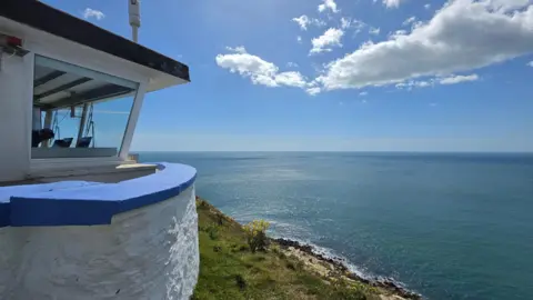 BBC Weather Watcher Susan Ward Worth Matravers, Dorset National Coastwatch Station - blue and white building to the left with steep grassy bank and sea stretching below.