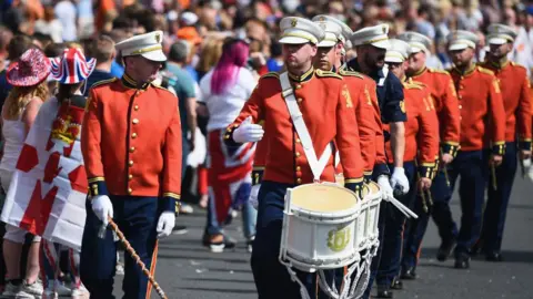 Getty Images A band marching wearing red uniforms and holding white drums.