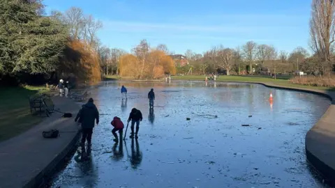 A frozen park pond with several young people walking and playing on the ice, surrounded by bare winter trees and a paved path under a bright blue sky.