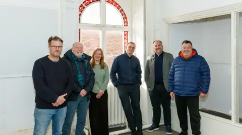 Neil Moore Six people - five men and one woman - stand in a row inside an empty room. The room is painted white and they are standing in front of a window. They are all smiling at the camera. 