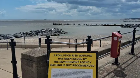 A sign reads "A pollution risk warning has been issues by the Environment Agency bathing is not recommended.

In the background is the sea with sand, rocks and sea defences visible. 
