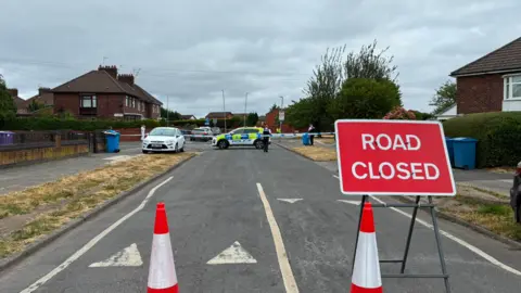 BBC Image shows cones and a 'Road Closed' sign blocking a residential street in Huyton, Merseyside. 
