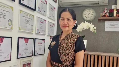 Real Thai Massage Maneerat Bossog standing by a wall of awards she has received. Her hair is in a low bun and she is smiling at the camera while wearing black and floral printed traditional wear.