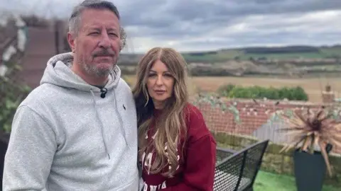 Heidi Tomlinson/BBC A man and woman standing in their garden with the quarry site in the background 