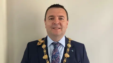 Derry mayor Ruairí Mc Hugh looking directly at the camera and smiling. He has very short, dark hair and is wearing a navy suit, pale blue shirt, navy patterned tie and his gold chain of office.