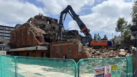 A large digger pulls down a big brick building