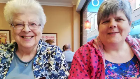Supplied Angela Lennox (on the right) and her mother sitting in a cafe and smiling. Miss Lennox's mother has short white hair and glasses and is wearing a blue jacket with flowers on it. Miss Lennox has short grey hair and is wearing a pink jacket with purple hearts on it.
