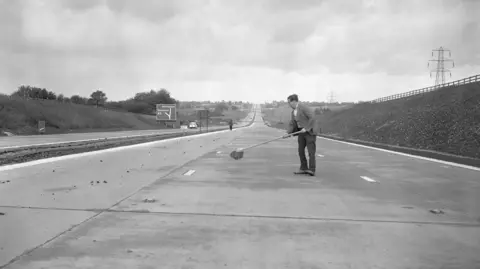 PA Media A black and white photo of a man holding a brush cleaning part of a newly construction motorway, dressed in an unexpectedly smart jacket