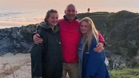 A man and two girls stand on the side of a cliff, which overlooks the beach and the sea.