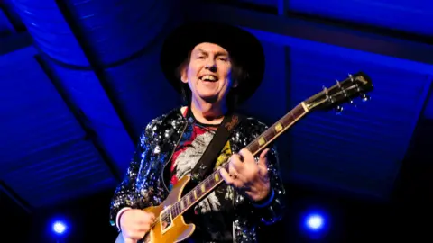 Getty Images A man wearing a cowboy hat and sparkly jacket on stage with a brown wooden-panelled guitar