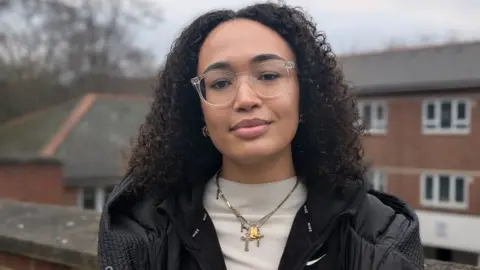 BBC Olimata stands outside in front of the camera and has long, curly black hair and glasses, wearing a black jacket, white top and neck jewelery