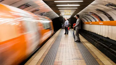 Glasgow subway station with the subway train blurred as it enters the station on the left. Two people stand on the platform.
