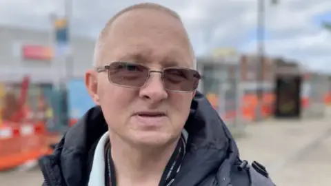 A man wearing tinted glasses looking into the camera. He is stood outside.