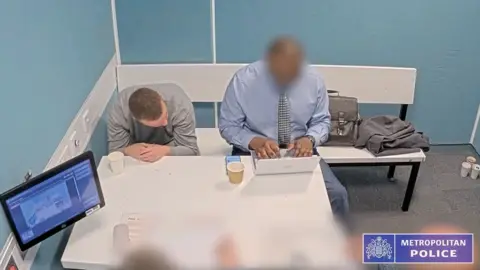 Man in grey sweater looks down to his right he is in a police custody room and man next t him, face blurred is typing on laptop