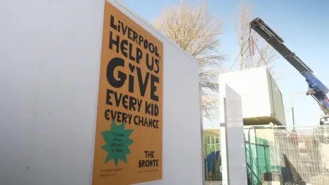 A yellow sign on a wall next to a building site which reads - Liverpool help us, give every kid every chance 