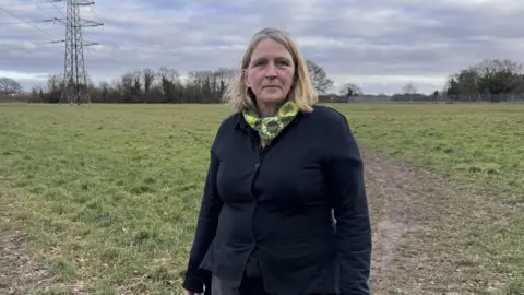 Colchester City Council Andrea Luxford Vaughan is a blonde woman wearing a navy cardigan and a green scarf. She is standing on the Middlewick site and looking at the camera.