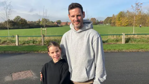 Steve Jones/BBC A young girl with brown hair stands next to her father, who also has dark hair, with a grass field in the background.
