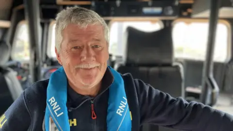 Close up of Jonathon Blakeston on the lifeboat. He is smiling at the camera. Mr Blakeston has a moustache and is wearing a navy fleece with a light blue RNLI life jacket over the top.