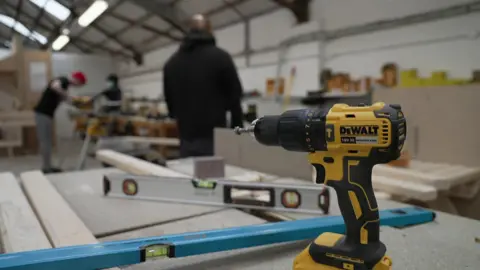 A drill and trade tools in focus with three anonymous male prisoners working in the background.