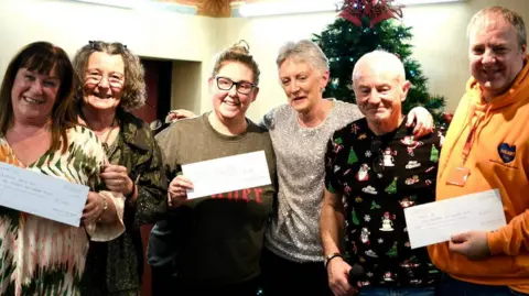 Six people, including Andrew Mears who is second from the right, are standing in a line with their arms slung over each others' shoulders and are smiling at the camera. Three are holding up large cheques. There is a Christmas tree behind them.