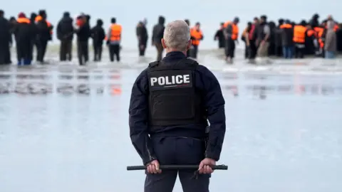 A police officer stands on a beach in Gravelands, France, with his back to the camera, wearing a black protective vest. He is watching as a group of people wearing orange life jackets enter the sea.