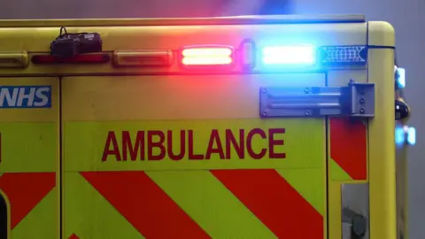 Close up of the back of an ambulance. The vehicle is yellow and has its orange / red and blue lights on. It has an NHS logo on the left back door and a camera fitted above the door. 