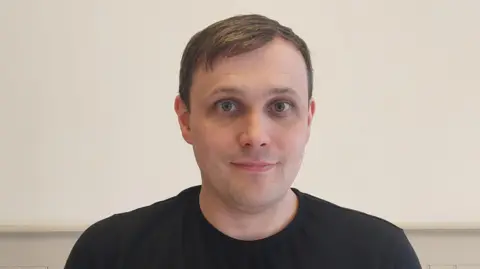 Darren Murphy has short brown hair and is wearing a black T-shirt. He is standing against a light-colored wall with wooden paneling.