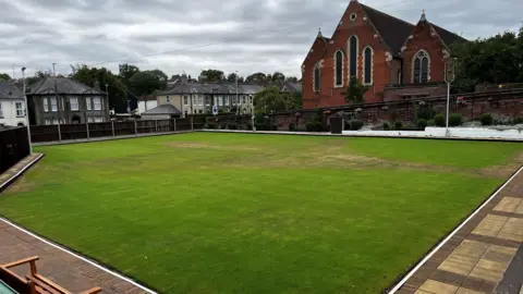 Jack Maclean/BBC A bowling green. Some of the grass is parched and brown. A bench is to one side of the green A brick wall and a wooden fence run round the perimeter of the area. A large church is next door to the green. Housing is opposite.