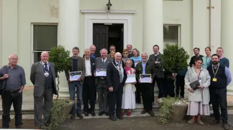 A group photograph showing the award winners and council dignitaries holding their certificates, standing outside a white building which has pillars.