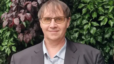 Mark Major A man in a grey shirt and grey suit, with brown hair and glasses stands infront of a wall with plants behind him. 